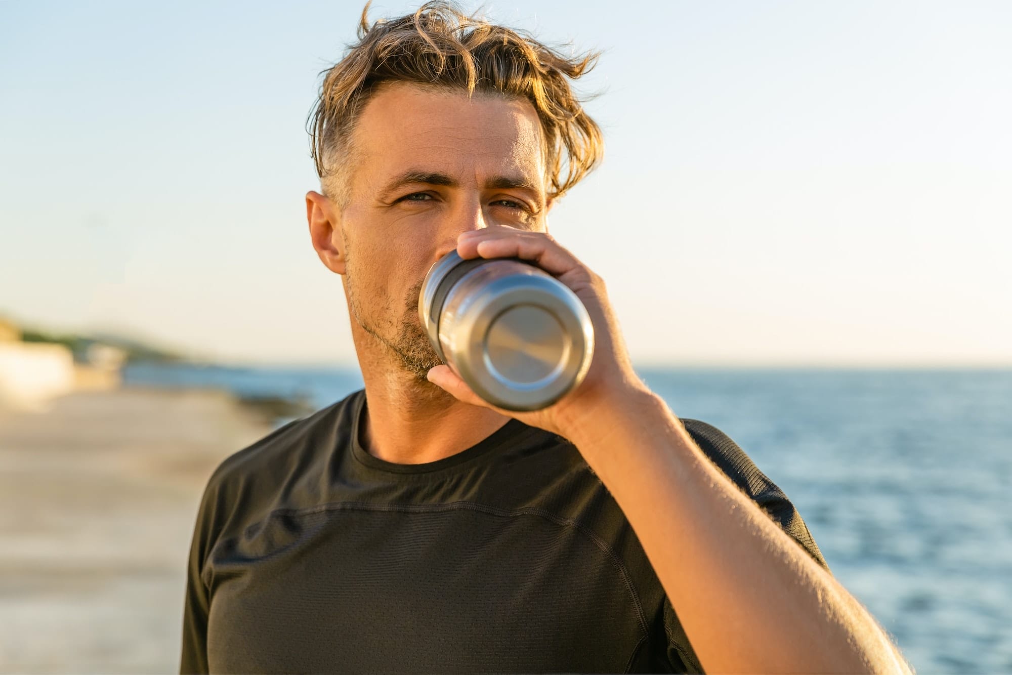 Handsome Adult Man Drinking Water from Fitness Bottle on Seashore in Front of Sunrise After Training
